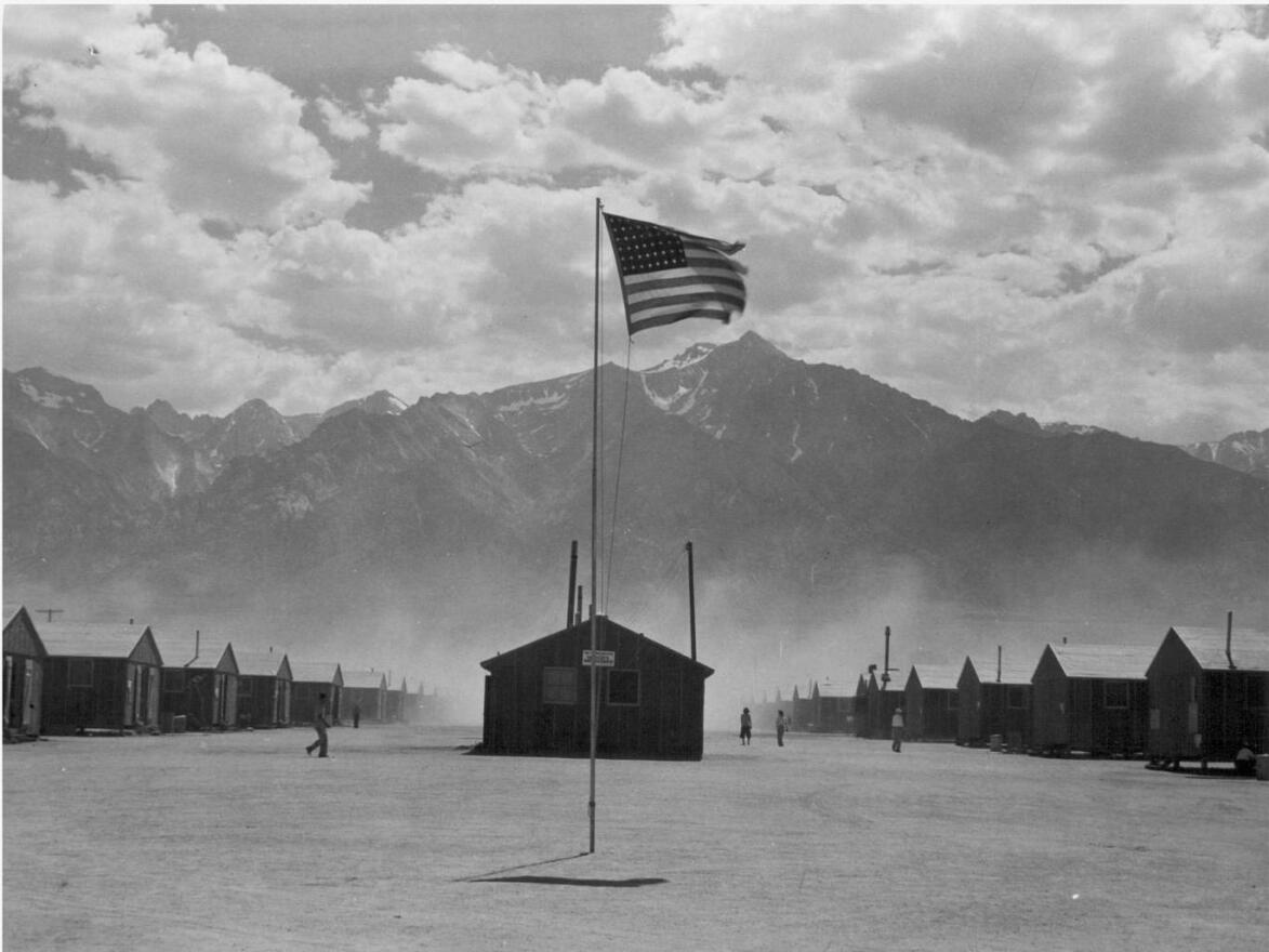 Scene of barrack homes at this War Relocation Authority Center for evacuees of Japanese ancestry. A hot windstorm brings dust from the surrounding desert. Photographer: Lange, Dorothea. Manzanar, Calif.